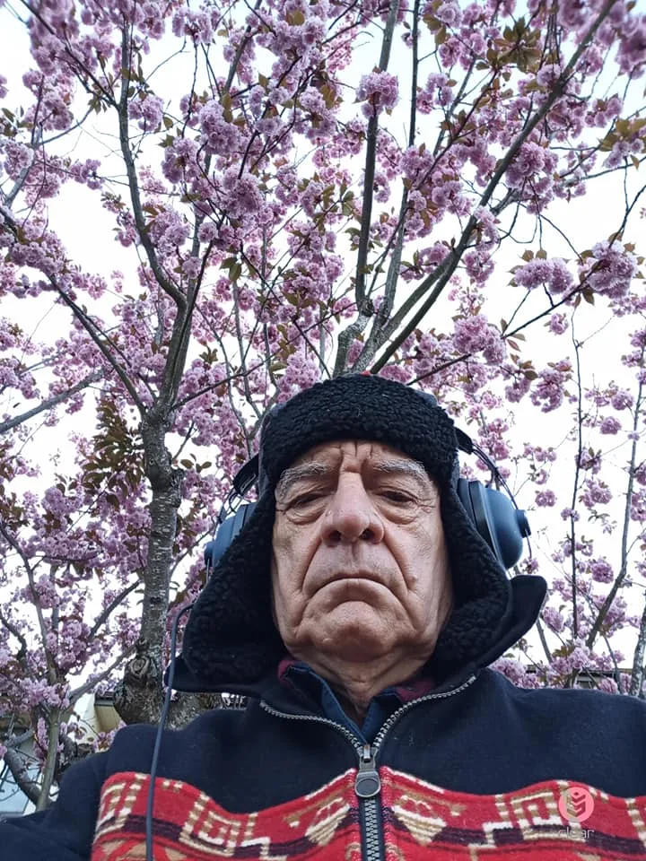 An elder in a black fleece trapper hat, headphones and a jacket with a red geometric pattern, looking down beneath a pink cherry tree in bloom.