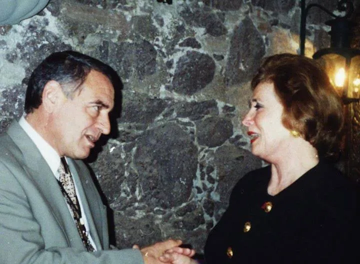 Phil Lane Jr. in a grey suit clasps hands and speaks with Jehan Sadat, who wears a dark jacket with gold buttons, before a stone wall lit by a lantern.