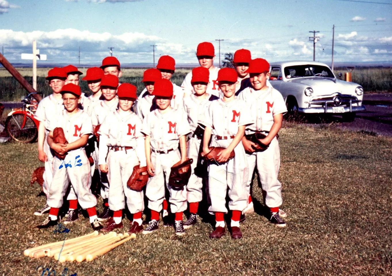 Vintage color photo of a boys' Little League team in white uniforms with red M letters, red caps and socks, holding gloves on grass beside bats; one boy is marked "ME" by hand.