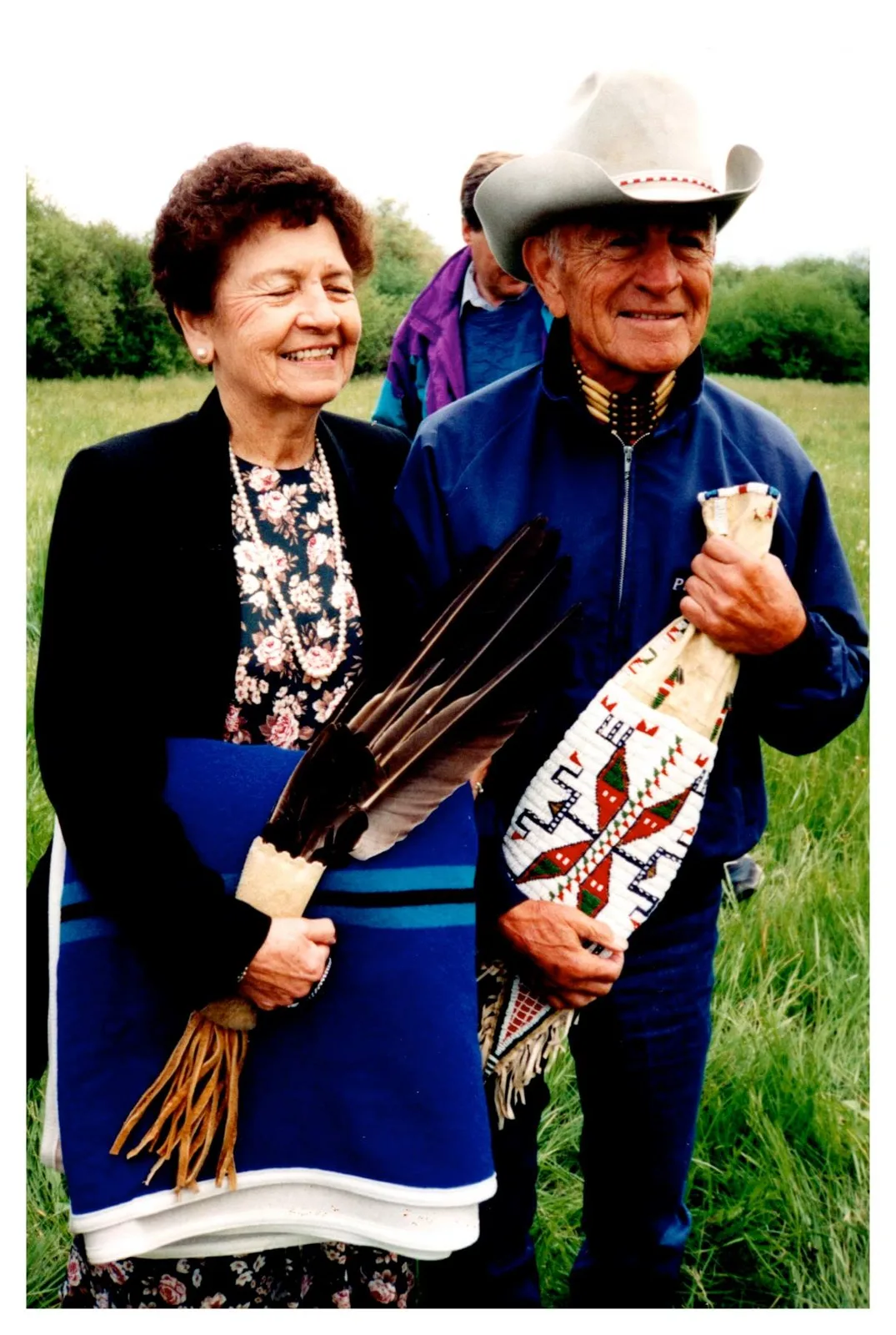 An elderly smiling couple stands in a grassy field. She holds a folded blue blanket and eagle feathers; he wears a cowboy hat and blue jacket and holds a beaded bag.