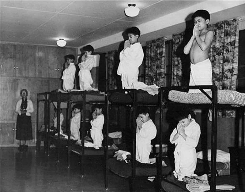 Black-and-white historical photograph of Indigenous boys in nightshirts kneeling in prayer on rows of bunk beds in a dormitory, watched by a woman standing at the left.