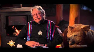 Dr. Dave Courchene Jr., an elder with grey hair wearing a dark ribbon shirt, seated by a wood stove beside a bear hide.