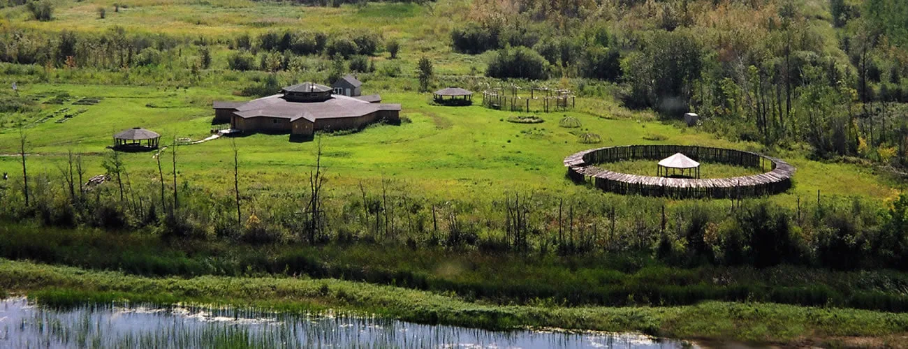 Aerial view of the Turtle Lodge, a round building in a green meadow near water, with gazebos and a circular log-fenced ceremonial enclosure.