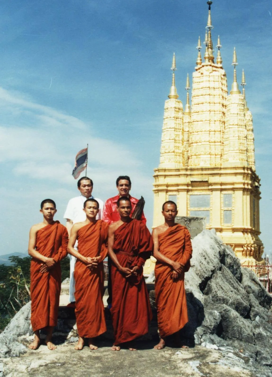 Four Buddhist monks in orange robes stand barefoot on a rocky outcrop, with a man in white and a man in a red shirt behind them, beside a golden temple spire and a Thai flag.