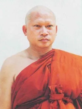 Portrait of a monk with a shaved head wearing an orange robe, standing against a pale wall.
