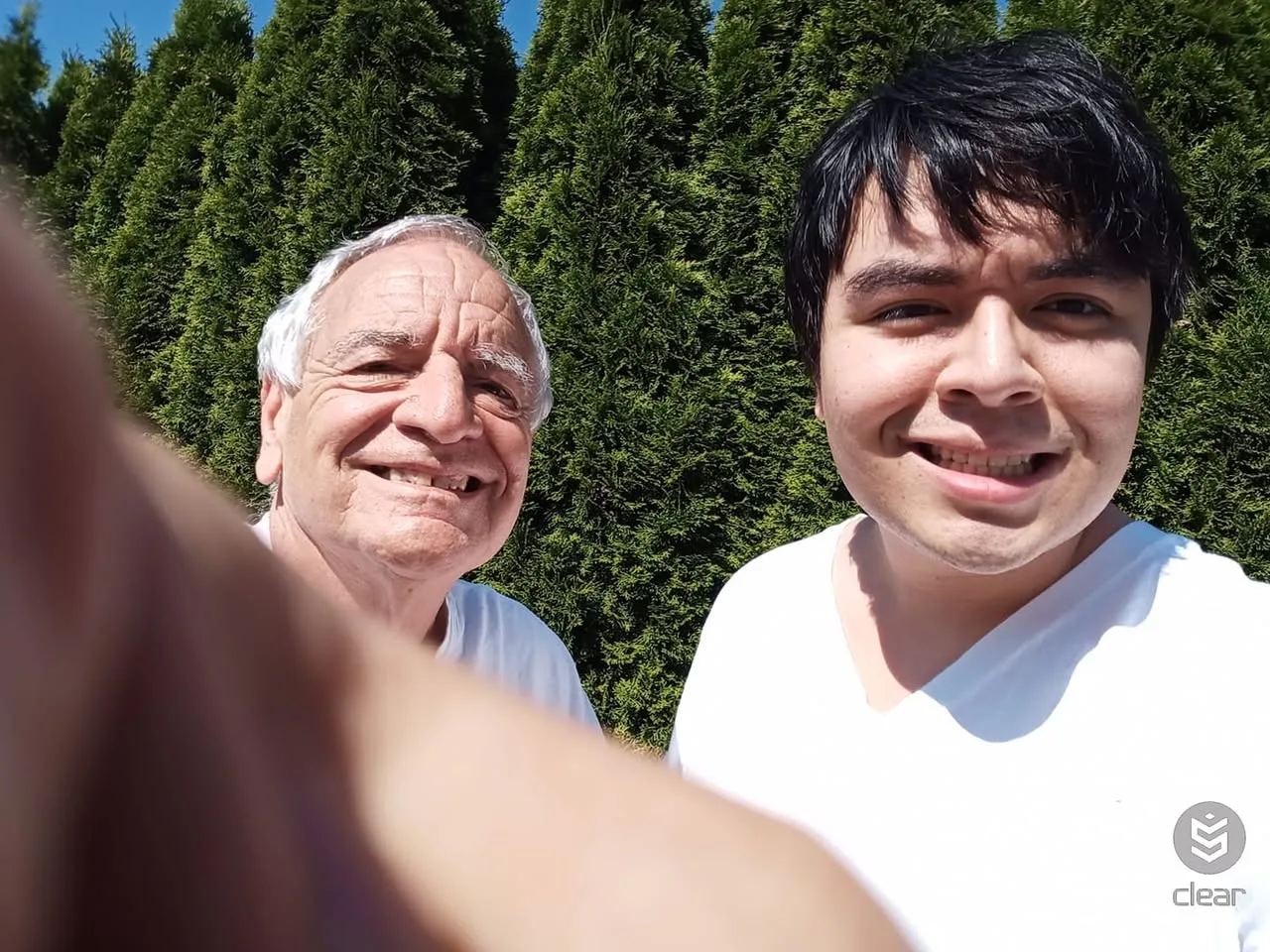A smiling white-haired elder and a smiling young man with dark hair, both in white T-shirts, take a sunny selfie in front of tall evergreen trees.