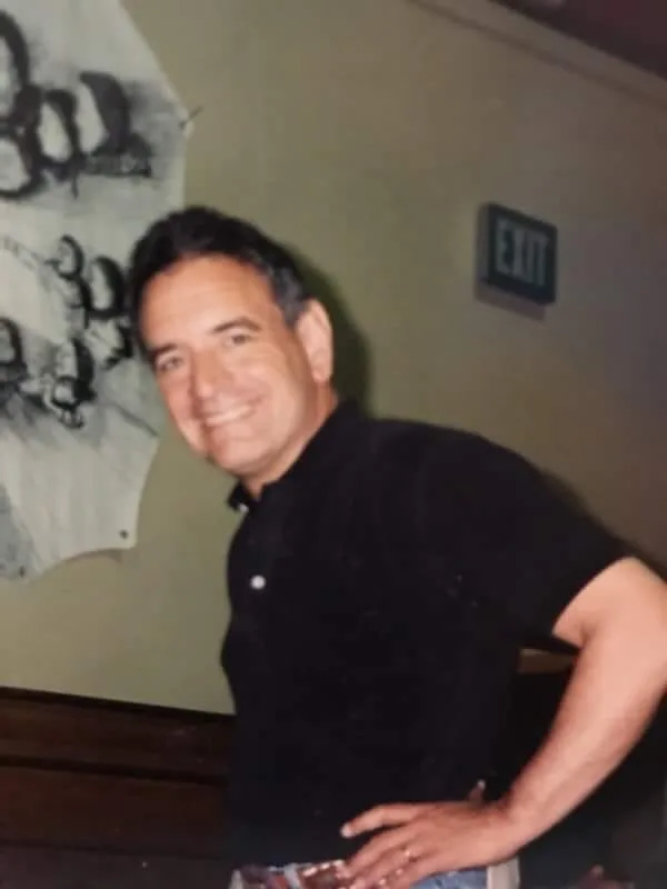 A smiling man with dark hair, in a black short-sleeved shirt, stands with his hand on his hip before a wall with a hanging drawing and an exit sign.