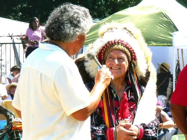 A man with gray curly hair holds a microphone to Phil Lane Jr., who smiles in a feathered headdress at the 2009 Seafair Pow Wow, with tents and onlookers behind.
