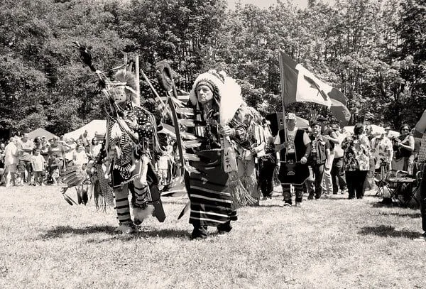 Black-and-white view of Phil Lane Jr. in a feathered headdress leading the Grand Entry with dancers in regalia, flags raised and spectators lining the field.