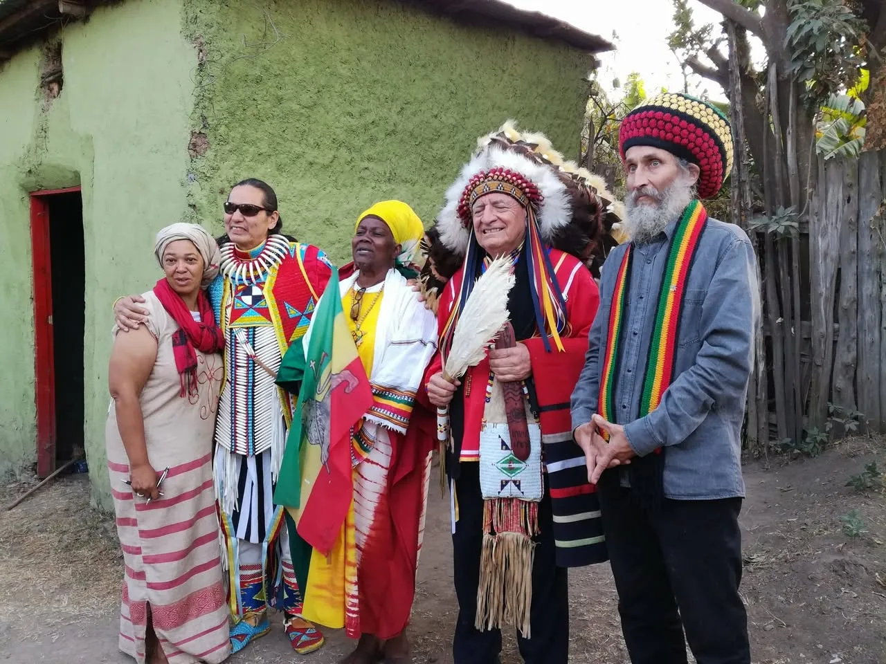 Five people stand together outside a green mud-walled house: a woman in a headscarf, a man in beaded regalia, a woman in yellow holding an Ethiopian flag, an elder in a feather headdress, and a bearded man in a Rasta hat.