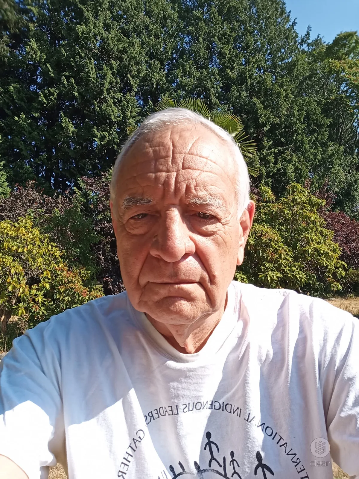 An elderly man with white hair takes a selfie outdoors in bright sun, wearing a white International Indigenous Leadership Gathering T-shirt, with tall evergreens behind him.