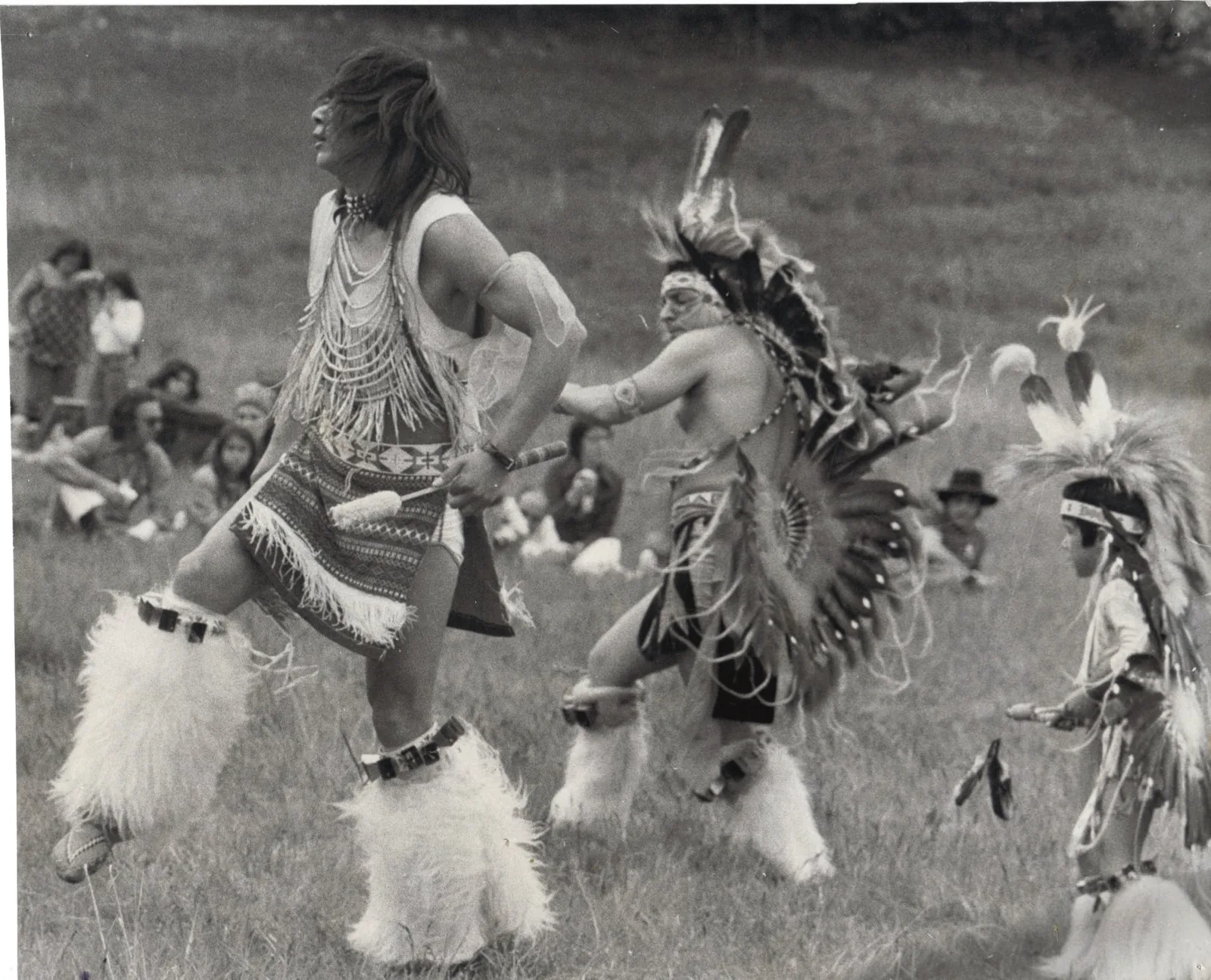 Black-and-white photo of three dancers in regalia with feather bustles, roaches and white fur leg wraps dancing on a grassy field, including a young boy, as people sit watching behind them.