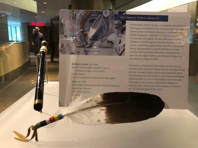 Museum display case holding a beaded bald eagle feather and a black lacquered flute, with a placard titled "Star Dancer: A Hero Among Us" about Commander John Herrington's space flight.