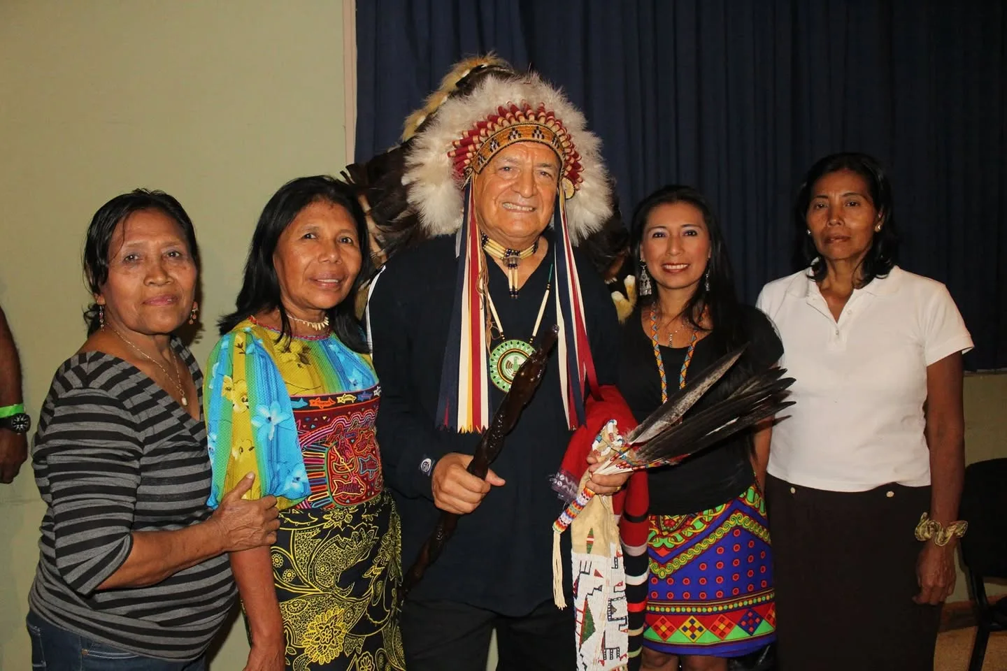 Phil Lane Jr. in a feathered headdress, holding a carved staff and eagle feathers, stands smiling with four Indigenous women, two in colorful traditional mola and patterned skirts.