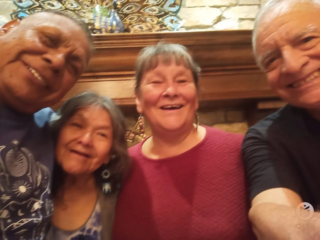 Four smiling elders pose close together for a selfie in front of a wooden fireplace mantel and stone wall, one wearing beaded earrings and another a red sweater.