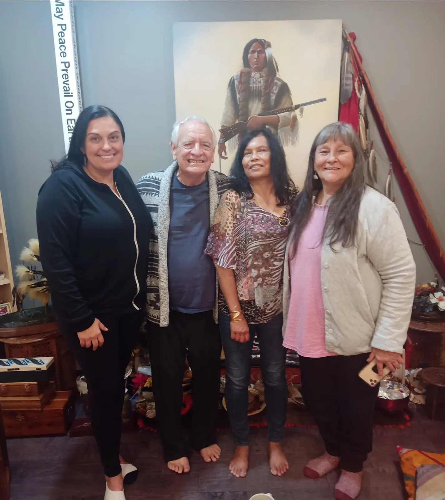 Four people stand barefoot and smiling together in a room before a painting of a warrior holding a rifle, beside a "May Peace Prevail On Earth" pole, feathers and a red staff.