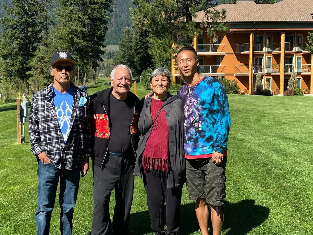 Four people stand together on a sunny lawn in front of a wooden lodge with pine trees and mountains behind: two older men, an older woman in a red top, and a younger man in a tie-dye sweatshirt.
