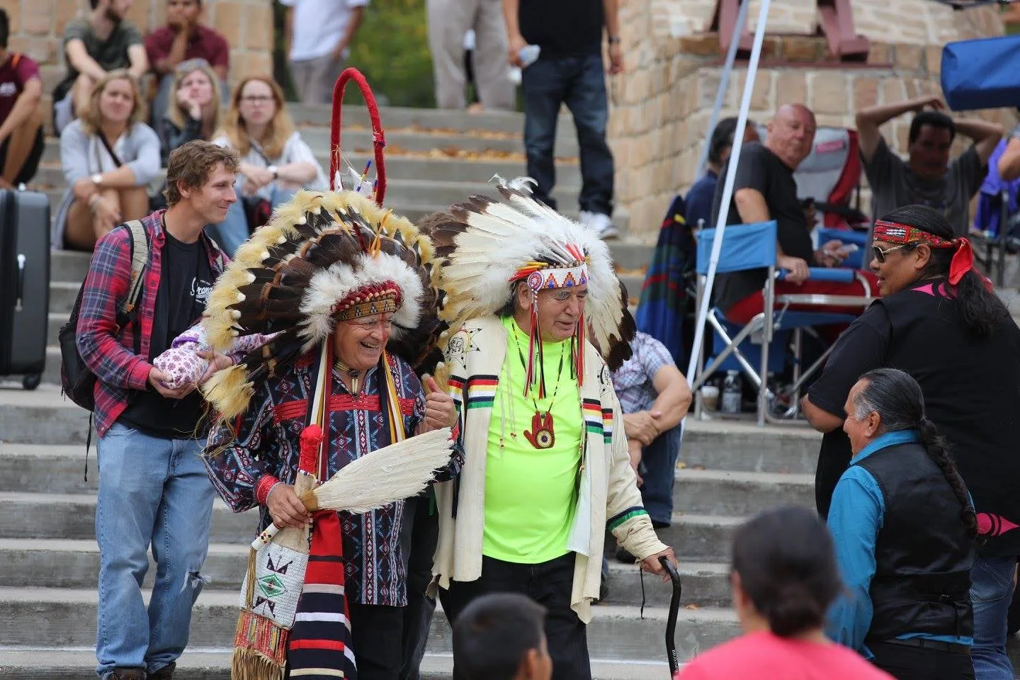 Two elders in eagle feather headdresses walk arm in arm, smiling, at an outdoor gathering in Winnipeg, one holding a feather fan and the other a cane, as people watch from stone steps.