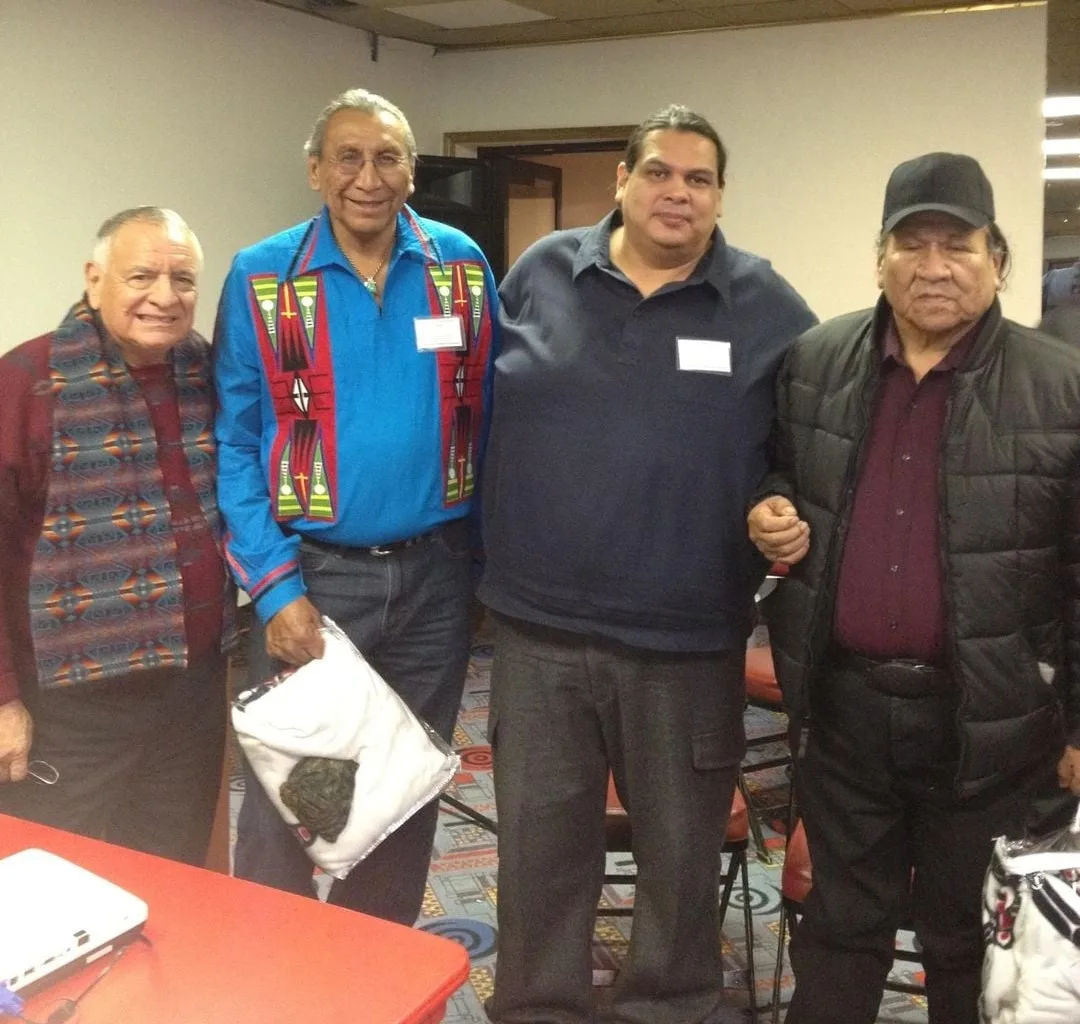 Four Indigenous men stand together in a meeting room. One wears a patterned blanket vest, another a blue ribbon shirt and holds a gift bag, and the eldest wears a black cap and jacket.