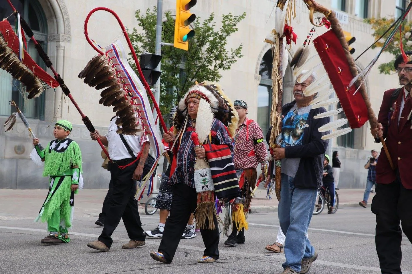 An elder in a feather headdress walks in a procession down a city street, flanked by people carrying eagle feather staffs and a child in green regalia.