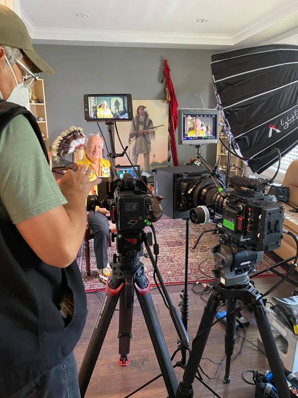 A film crew with two cinema cameras on tripods and a large softbox light records an elder in a yellow shirt, seated beside a feather headdress and a painting of an Indigenous warrior.
