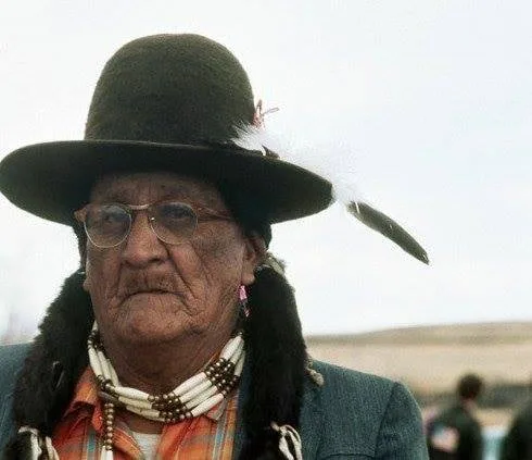 An elderly Lakota man in glasses and a dark brimmed hat with a white feather, wearing a bone choker and braid wraps, stands outdoors on the open prairie.