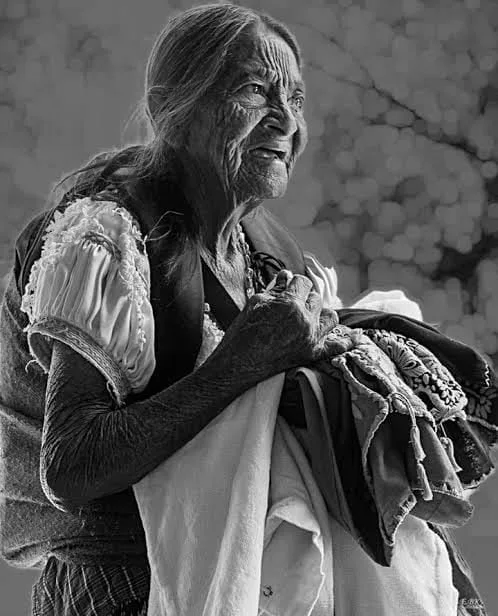 Black-and-white portrait of an elderly woman with a deeply lined face and long gray hair, wearing an embroidered blouse and holding folded cloths.