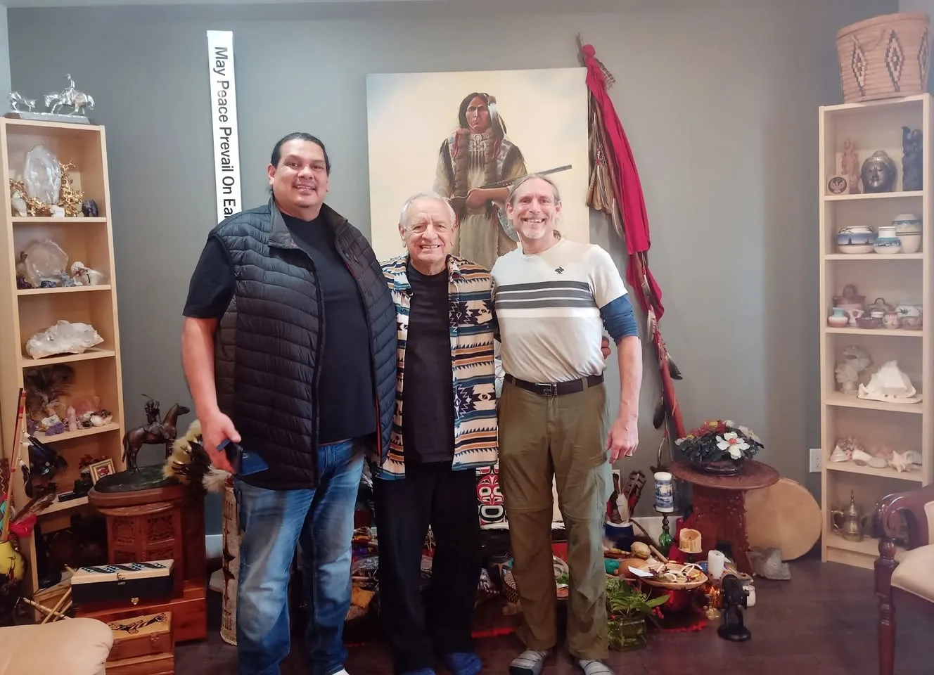 Three smiling men stand arm in arm in a room filled with Indigenous art, baskets, crystals, a drum and a feathered staff, beneath a painting of a warrior and a "May Peace Prevail On Earth" pole.