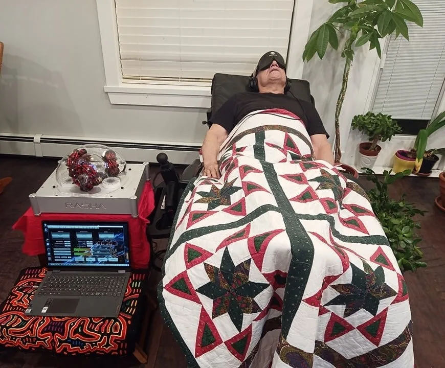 A person reclines in a black chair wearing headphones and an eye mask, covered by a red, green and white star quilt, beside a RASHA device with glowing coils and a laptop.