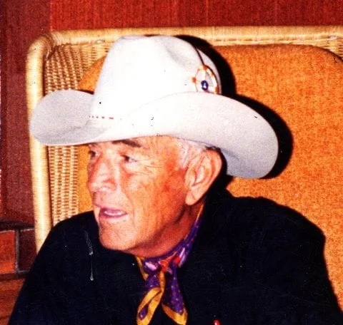 Phil Lane Sr., an elder in a white cowboy hat with a beaded medallion and a patterned neckerchief, seated in a wicker chair and smiling.