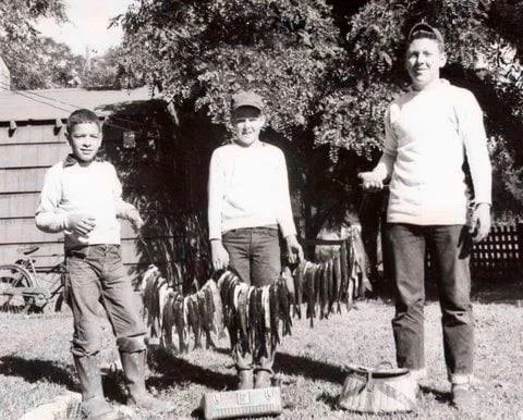 Black-and-white photo of three boys standing on a lawn beside a house, holding a long string of caught fish between them, with a bicycle and trees in the background.