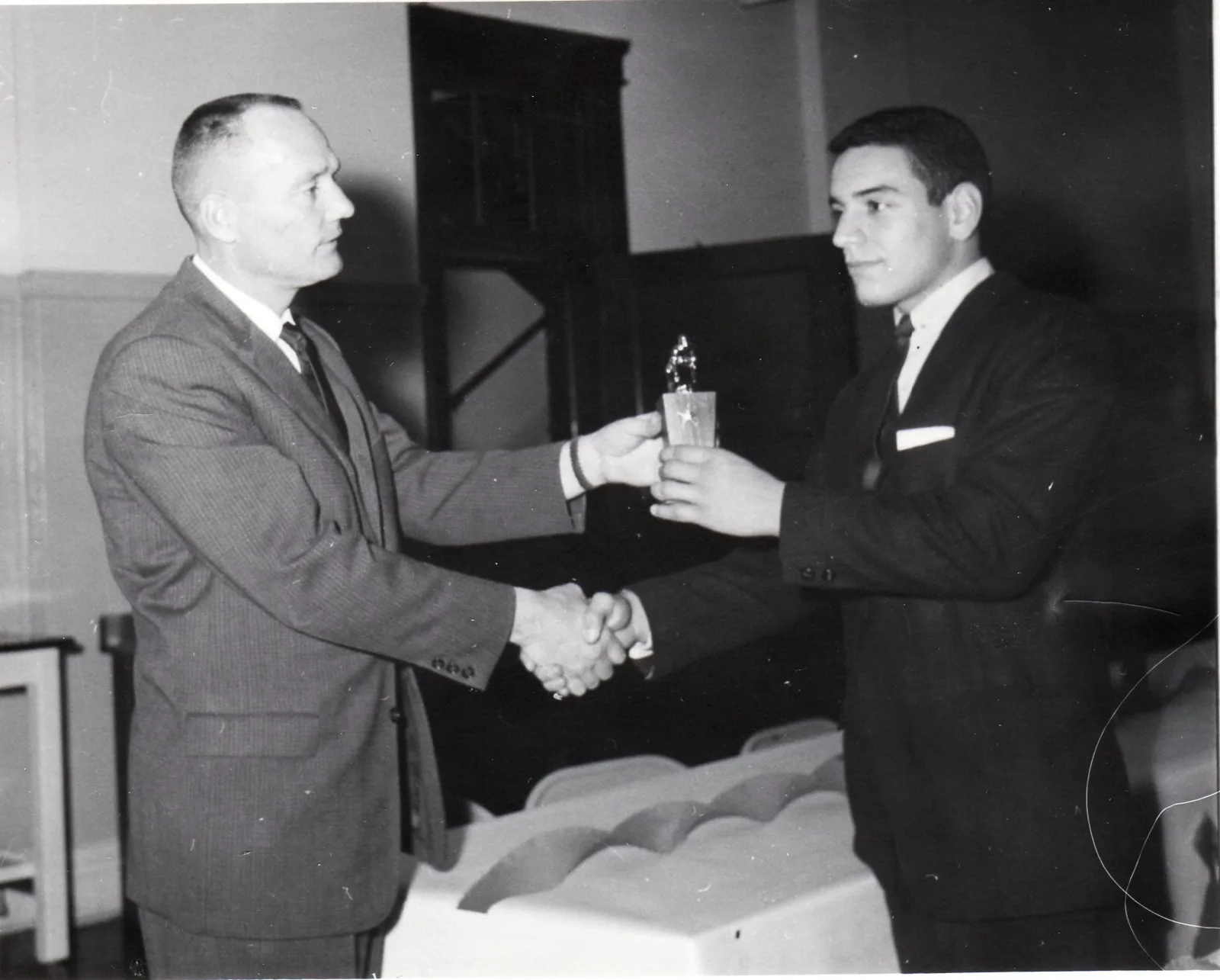 Black and white photo of an older man in a suit shaking hands with a young man in a dark suit while handing him a small trophy, in a hall with a white-draped table.