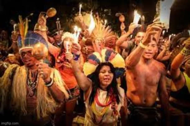 A night gathering of Indigenous people in feather headdresses, beads and body paint, raising candles and rattles as they chant together.