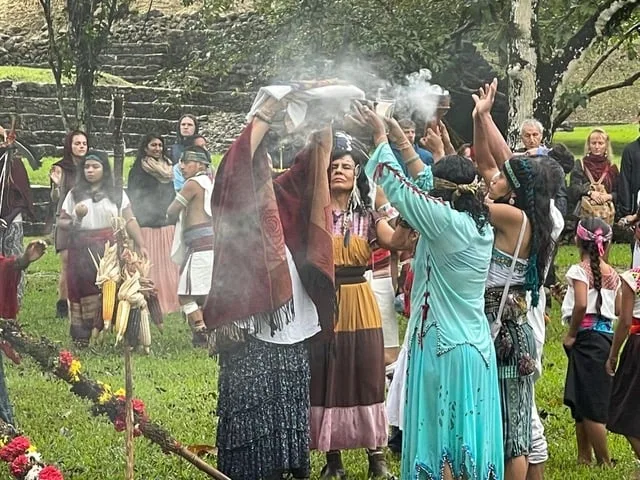 Women in traditional dress raise smoking incense and white cloth bundles skyward in a ceremony on the grass before stone ruins, with ears of corn and flowers nearby.