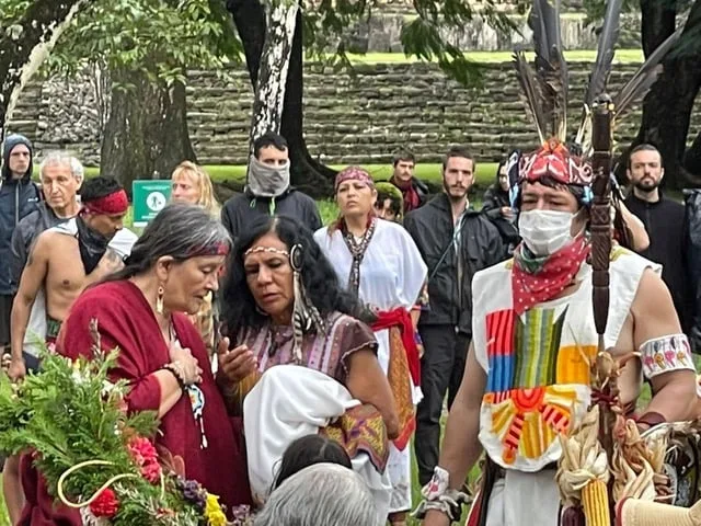 Elder women in red and embroidered garments stand beside a man in a feathered headdress holding a feathered staff, with onlookers and stone ruins behind them.