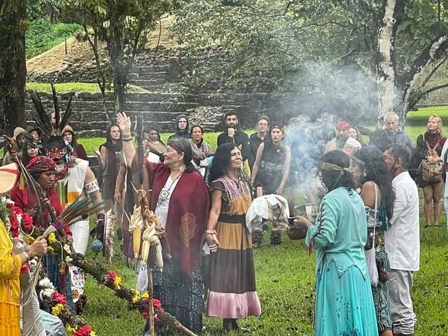 A circle of people in traditional clothing gathers in ceremony as incense smoke rises, with a feathered staff, corn and flowers before a terraced stone ruin.