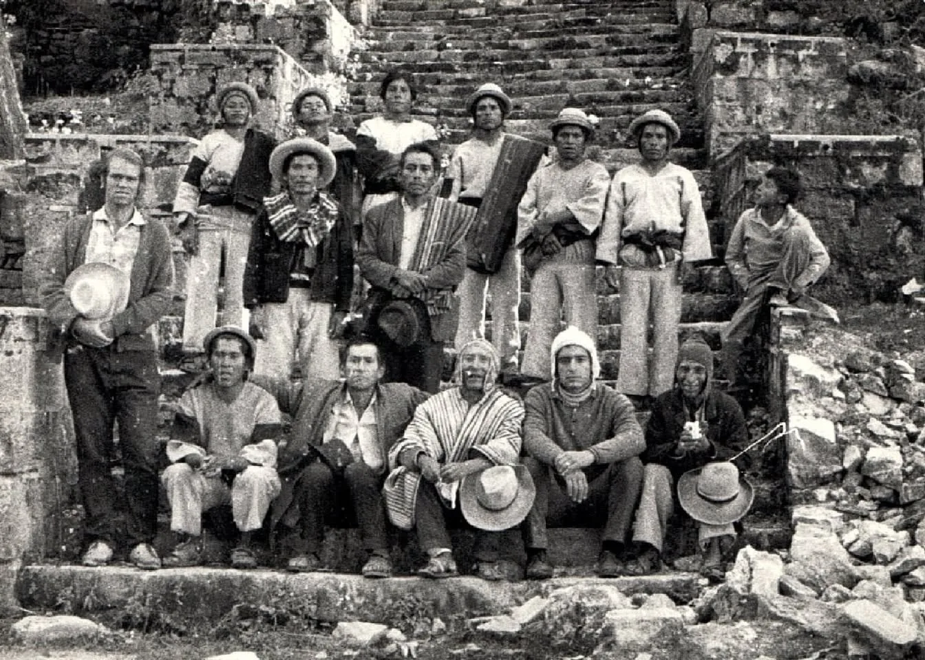 Black-and-white group photo of about sixteen men, many in woven ponchos and brimmed hats, seated and standing on weathered stone stairs.