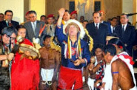 Indigenous spiritual leaders in traditional dress, one in a white feathered headdress with a raised fist, stand in a hall surrounded by men in suits.