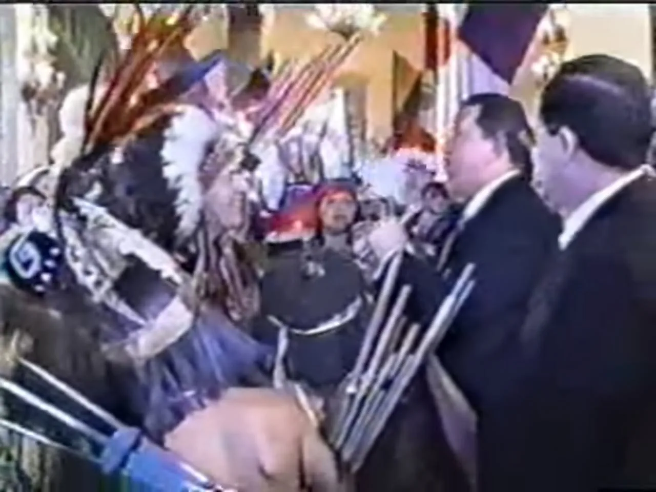 Blurry video still of a person in a large feathered headdress facing men in dark suits, with flags and a crowd in the background.