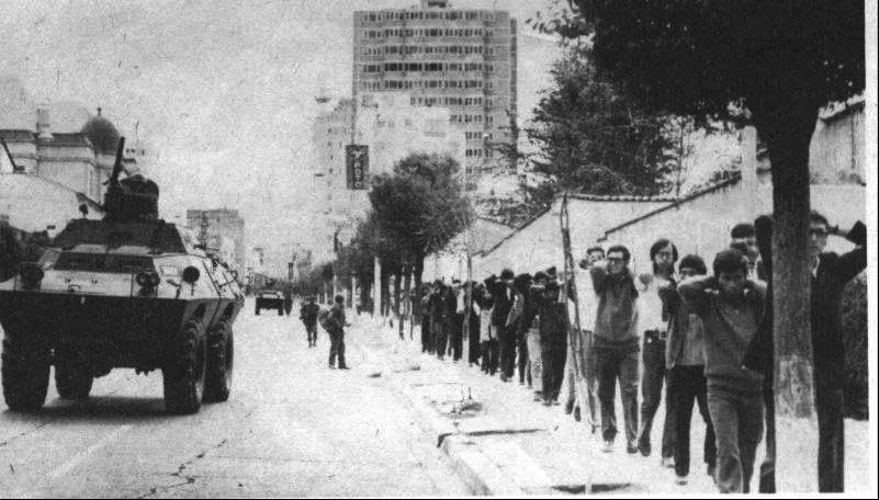 Black-and-white photo of a city street where an armored military vehicle sits as a long line of young men walk along the sidewalk with hands raised behind their heads, soldiers nearby.