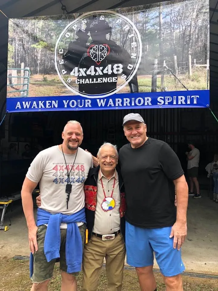 Phil Lane Jr., an elder wearing an eagle medallion, stands smiling between Adam Marr and Mark Rypien beneath a banner reading "4x4x48 Challenge: Awaken Your Warrior Spirit."