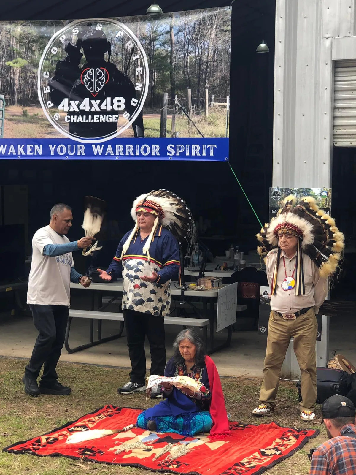 A man in a white T-shirt smudges with an eagle feather fan as a man in a feather headdress holds out his hands in prayer. An elder woman sits on a red blanket with a pipe bag, and a second elder in a headdress stands nearby. Behind them, a banner reads "4x4x48 Challenge: Awaken Your Warrior Spirit."