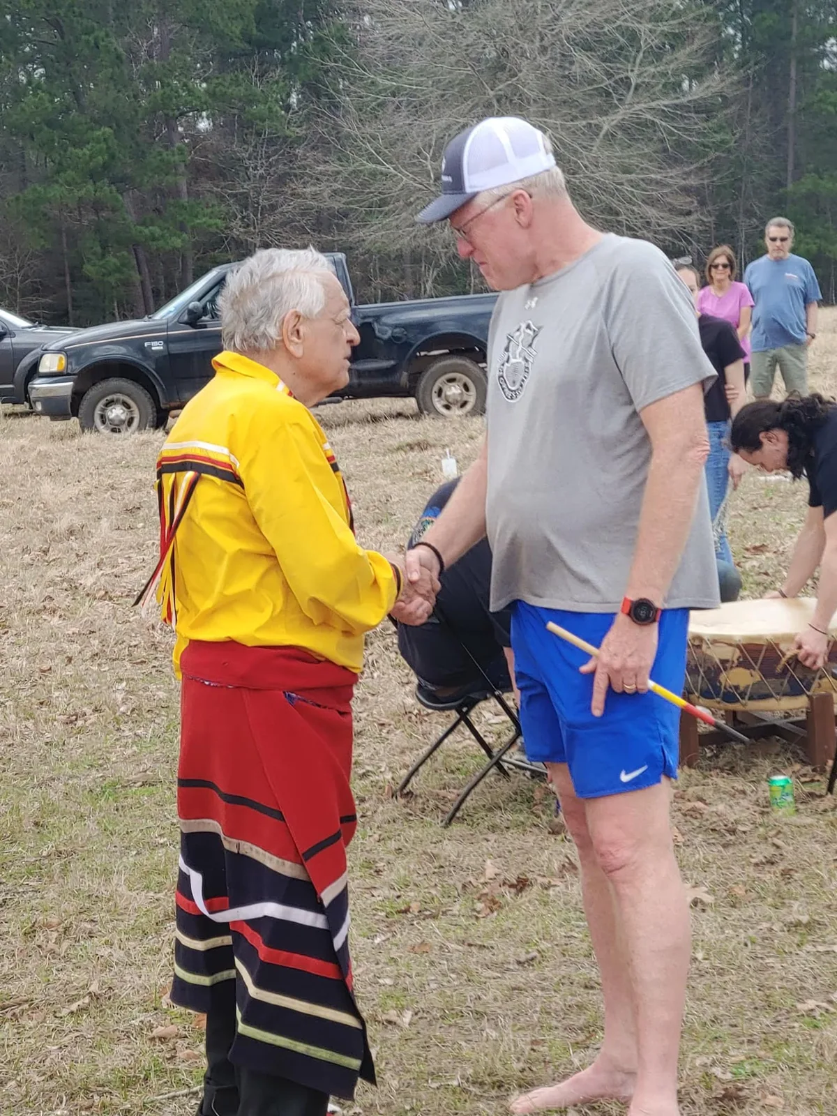 An elder in a yellow ribbon shirt and red and black striped skirt shakes hands with a tall barefoot man in a cap and gray T-shirt in a field, a drum and onlookers behind them.