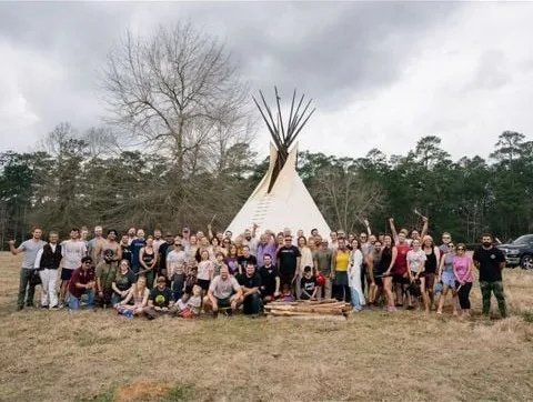 A large group of people standing, kneeling and sitting in a grassy field in front of a white tipi, with bare trees and forest under a cloudy sky; some raise their arms.