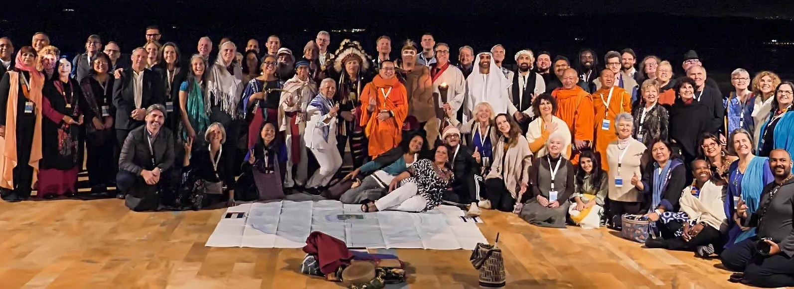 A large interfaith group poses at night on a wooden floor by the Dead Sea, including an elder in a feather headdress, monks in orange robes and a man in white Gulf dress.