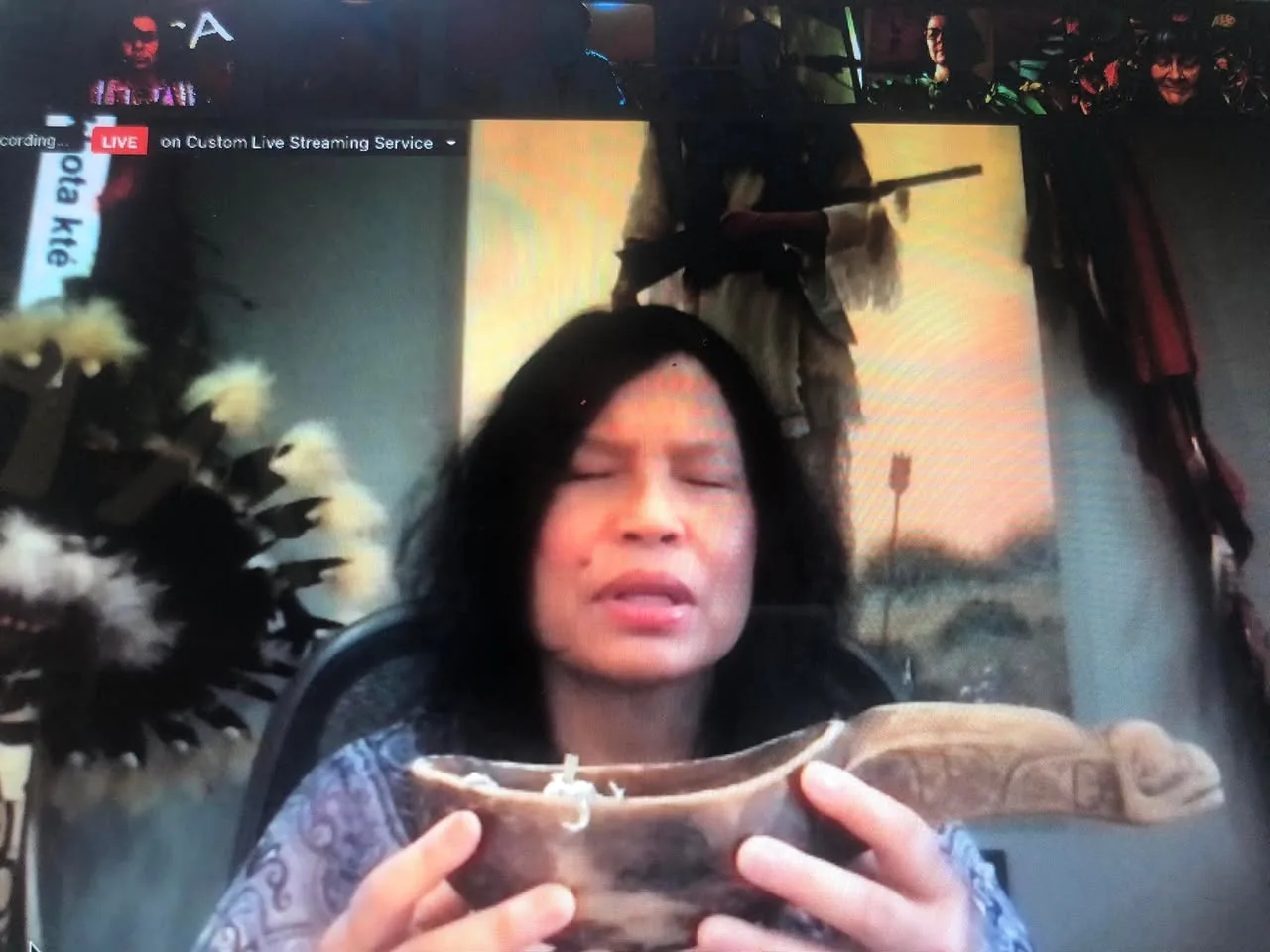 Screenshot of a livestreamed video call: a woman with long dark hair, eyes closed in prayer, holds a carved wooden ceremonial cup, with a feather headdress beside her.
