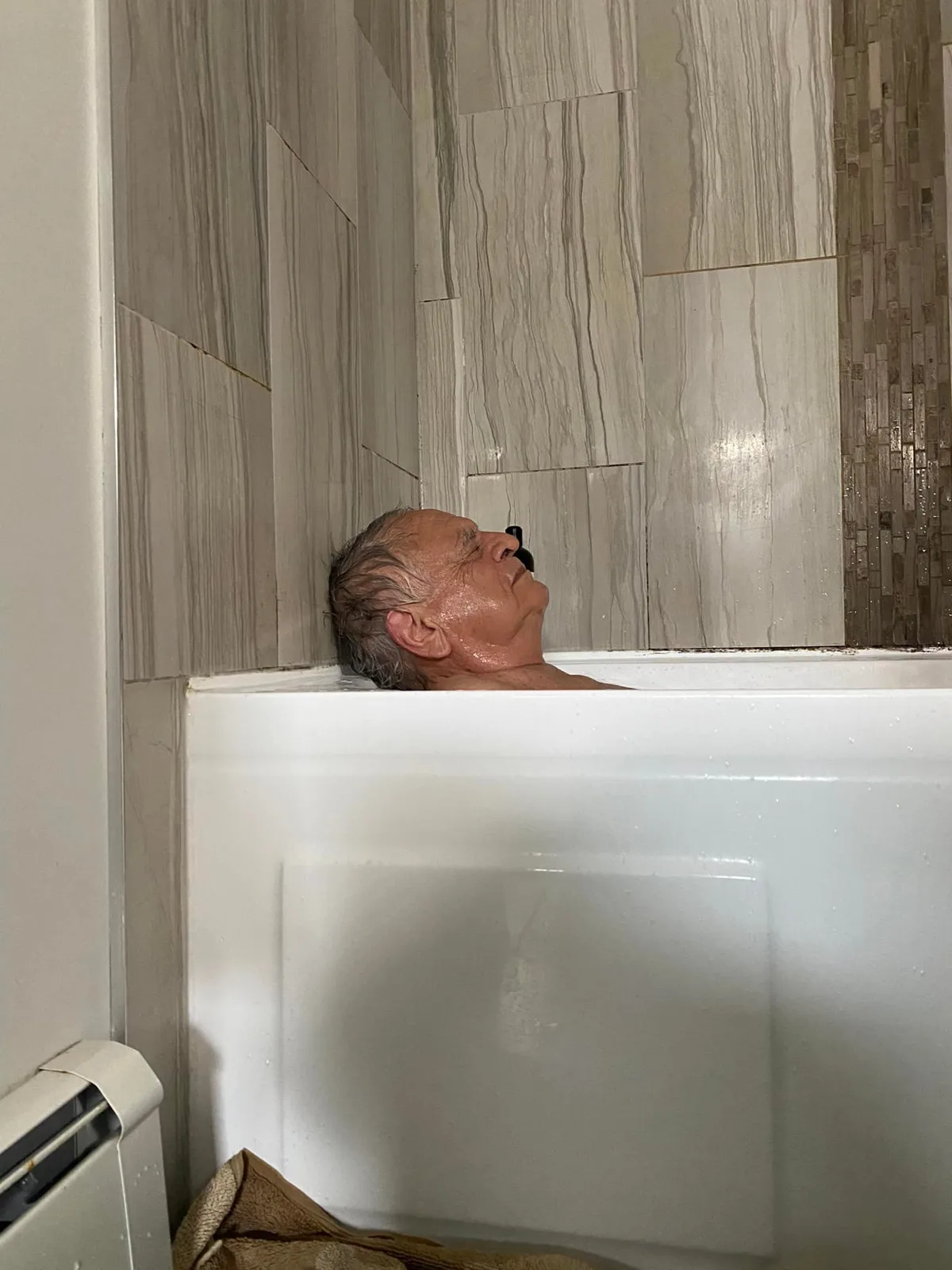 An older man with wet gray hair reclines with eyes closed in a white bathtub, submerged to his shoulders, against gray marbled tile walls.