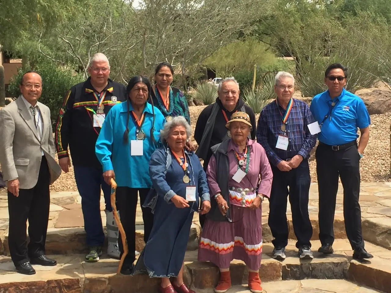 Group of ten Indigenous elders and leaders standing on stone steps in a desert garden, many wearing medallions on ribbons, name badges and traditional dress and jewelry.