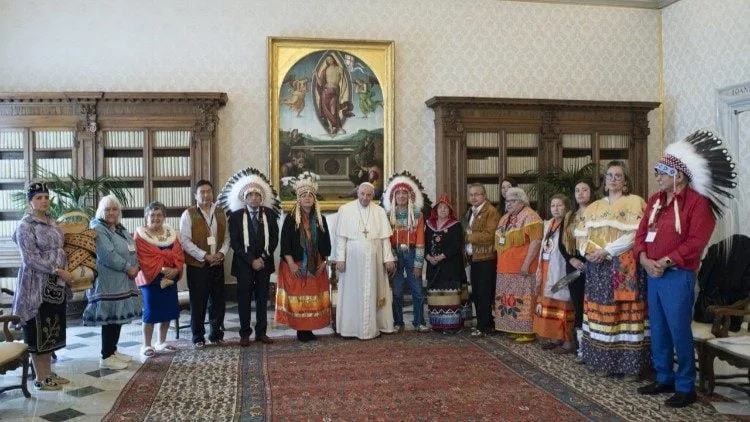 The Pope in white robes stands with a delegation of Indigenous people, some in feather headdresses and ribbon skirts, in a Vatican room beneath a large religious painting.