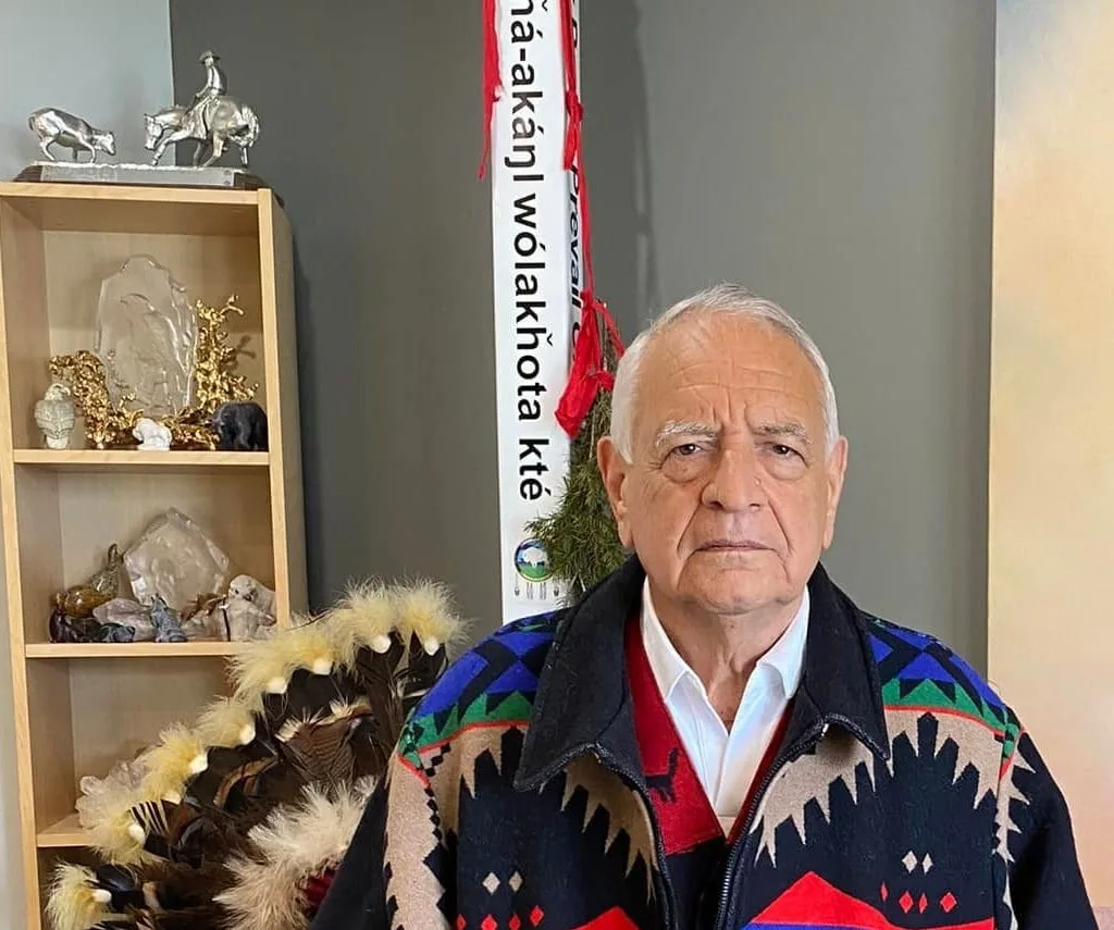 An elderly man with white hair in a colorful Native-patterned jacket sits before a feathered staff, a shelf of carvings, and a white banner with Lakota words and red ties.